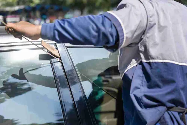 Someone in a blue work jacket holds a car door open at the top with a wedge and reaches inside with a long thin rod.
